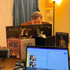 A photograph of Ben's head peeking above the wall of Steve Martin albums, books, and DVD sets he consulted for the episode, as seen from the vantage of Pete's recording station.